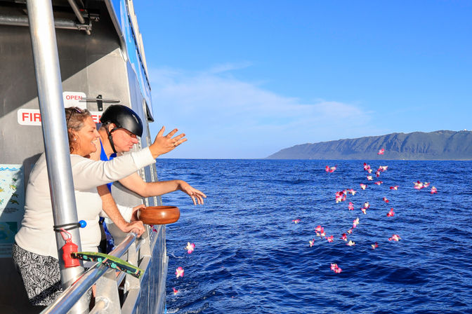 Two passengers on a boat scatter pink flower petals into the deep-blue open ocean toward a distant rugged island coastline under a bright, clear sky.