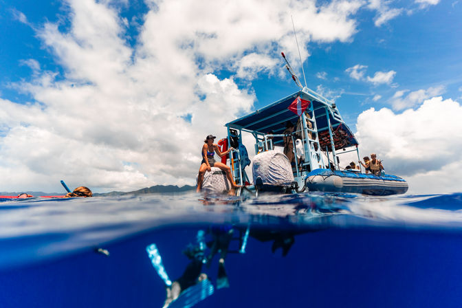 Split-view of snorkelers and scuba divers underwater with a blue dive boat and passengers above in clear tropical ocean near a distant island under a partly cloudy sky