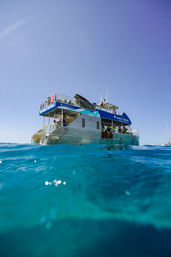 Two-level tour catamaran floating in turquoise tropical ocean under a clear blue sky, passengers on deck enjoying a sunny day