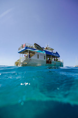 Two-level tour catamaran floating in turquoise tropical ocean under a clear blue sky, passengers on deck enjoying a sunny day