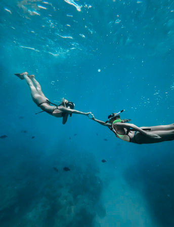 Two snorkelers holding hands as they glide over a turquoise tropical coral reef, underwater snorkeling scene with scattered fish and sunlit water