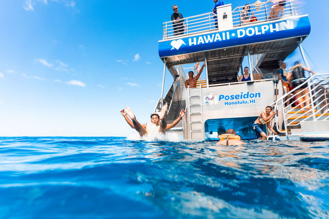 Passengers leaping from a sunny tour catamaran into crystal-blue Pacific waters off Honolulu, Hawaii, with crew and swimmers nearby.