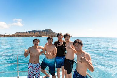 Five young men on a boat off Waikiki, Hawaii, posing with shaka signs and Diamond Head crater in the background over bright blue Pacific waters.