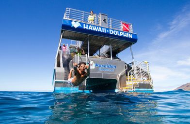 Person diving off a two-level snorkeling tour boat into the clear blue Hawaiian ocean near a distant island under a sunny sky.