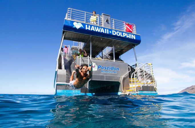 Person diving off a two-level snorkeling tour boat into the clear blue Hawaiian ocean near a distant island under a sunny sky.