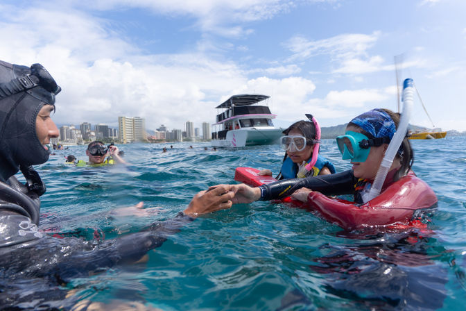 Two young snorkelers in masks and life jackets practicing with an instructor during a tropical boat snorkeling tour in clear turquoise water, catamaran and coastal city skyline in the background.