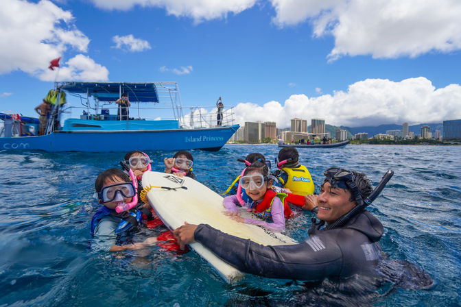 Smiling snorkel instructor guides a group of kids in life jackets holding a surfboard in the blue ocean, with a tour boat and coastal city skyline under a sunny, cloud-dotted sky.