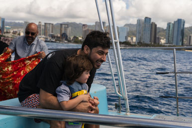 Man laughing as he holds a young child wearing a yellow life jacket on a boat, looking out over blue ocean with a coastal city skyline of tall buildings in the background.
