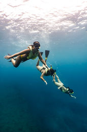 Three snorkelers in yellow vests and fins exploring clear blue tropical ocean above a coral reef, sunlight rippling on the water surface
