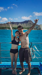 Happy young couple in swimwear on a boat, arms raised in shaka signs, turquoise Pacific waters with Diamond Head crater and Waikiki skyline in Honolulu, Hawaii.
