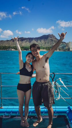Happy young couple in swimwear on a boat, arms raised in shaka signs, turquoise Pacific waters with Diamond Head crater and Waikiki skyline in Honolulu, Hawaii.
