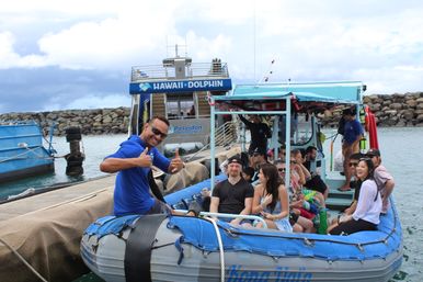 Smiling boat guide gives thumbs-up from an inflatable tour boat filled with laughing passengers at a dock beside a rocky breakwater in a coastal harbor.