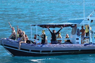 Snorkeling tour on an inflatable boat in turquoise tropical water, passengers in life jackets and snorkel gear smiling and waving under a shaded canopy