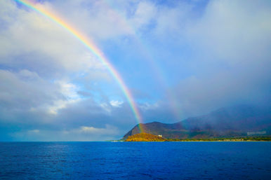 Vivid double rainbow arching from a cloudy sky to a sunlit tropical island coastline, with deep blue ocean and rugged green mountains