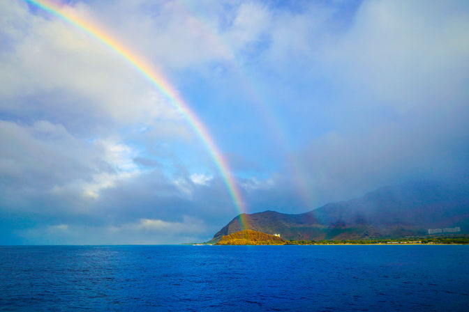 Vivid double rainbow arching from a cloudy sky to a sunlit tropical island coastline, with deep blue ocean and rugged green mountains