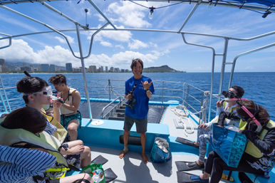 Group of snorkelers on a blue tour boat off Waikiki, Honolulu, with Diamond Head in the background; guide flashing a shaka as passengers put on masks, fins and life vests under sunny skies.