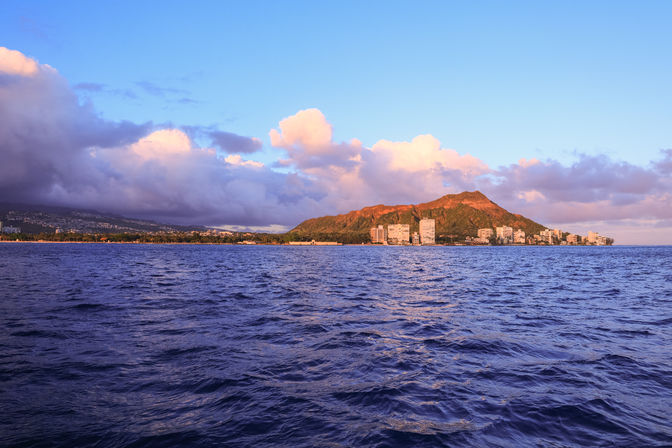 Vibrant sunset view of Diamond Head crater and Waikiki coastline from the ocean, purple-blue waves in the foreground and golden clouds overhead.