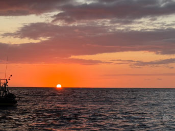Golden-orange sun dipping below the ocean horizon, rippling dark water and a small boat silhouette at left under a layered cloudy sky.