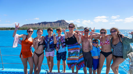 Group of people on a sunny Waikiki boat tour in Honolulu, posing with shaka signs against turquoise Pacific waters and Diamond Head crater.