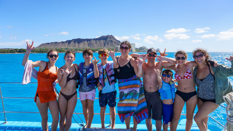 Group of people on a sunny Waikiki boat tour in Honolulu, posing with shaka signs against turquoise Pacific waters and Diamond Head crater.