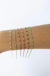 Close-up of a forearm wearing multiple delicate gold chain bracelets with tiny colorful beads (white, turquoise, blue, pink, black, green) stacked against a clean white background