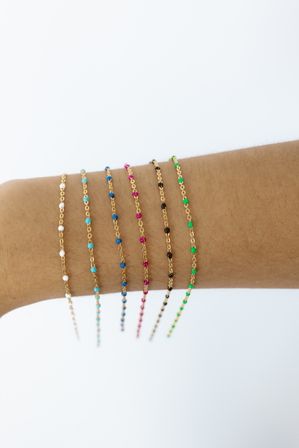 Close-up of a forearm wearing multiple delicate gold chain bracelets with tiny colorful beads (white, turquoise, blue, pink, black, green) stacked against a clean white background