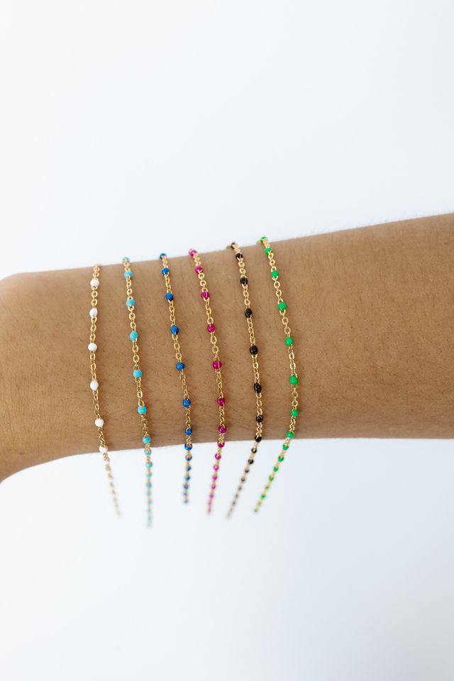 Close-up of a forearm wearing multiple delicate gold chain bracelets with tiny colorful beads (white, turquoise, blue, pink, black, green) stacked against a clean white background