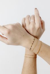 Close-up of crossed wrists wearing stacked delicate gold bracelets and a small black-beaded chain on a clean white background
