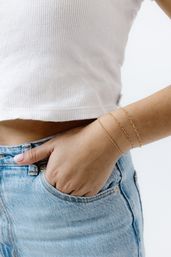 Close-up of a hand tucked into light-blue denim jeans pocket wearing three delicate gold chain bracelets and a white ribbed crop top