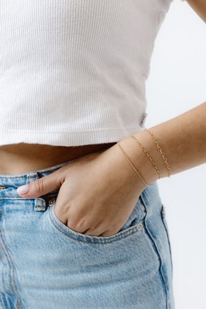 Close-up of a hand tucked into light-blue denim jeans pocket wearing three delicate gold chain bracelets and a white ribbed crop top