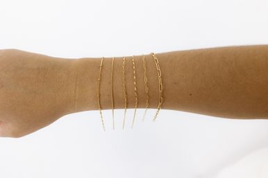 Close-up of a forearm wearing six dainty gold chain bracelets stacked along the wrist on a clean white background — minimalist jewelry wrist shot.