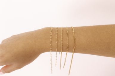 Close-up of a wrist wearing five dainty gold chain bracelets in different styles, stacked on a soft pink background