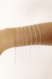 Close-up product shot of a wrist wearing seven delicate silver chain bracelets stacked in varying link styles — minimalist jewelry on a soft neutral background.