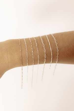 Close-up product shot of a wrist wearing seven delicate silver chain bracelets stacked in varying link styles — minimalist jewelry on a soft neutral background.