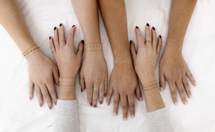 Overhead flat-lay of six diverse hands wearing delicate gold and silver chain bracelets and simple rings, posed on white linen — minimalist jewelry close-up.