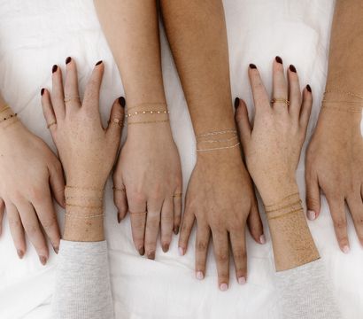 Overhead flat-lay of six diverse hands wearing delicate gold and silver chain bracelets and simple rings, posed on white linen — minimalist jewelry close-up.