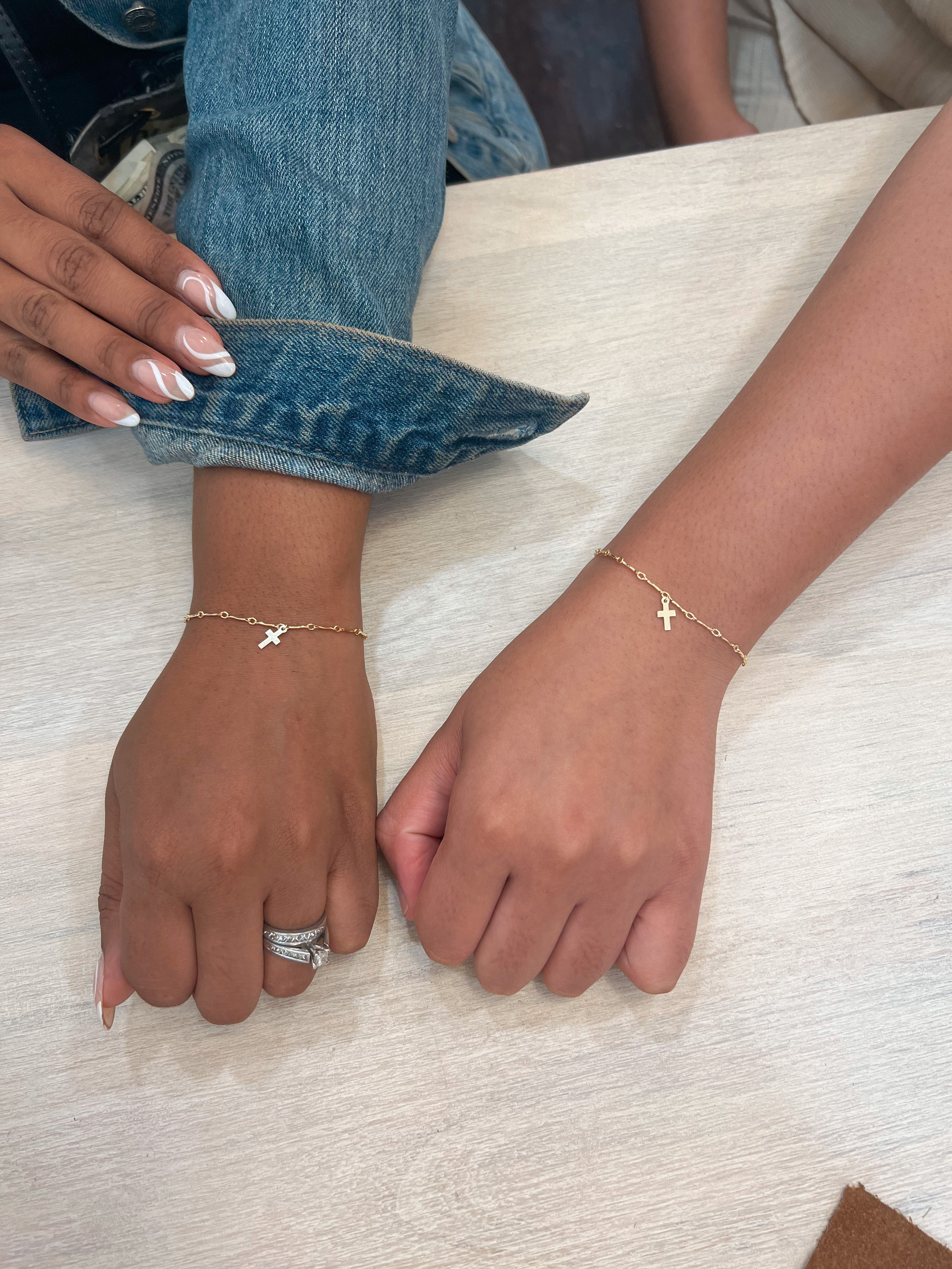 Two women's wrists on a light wood table wearing matching delicate gold chain bracelets with small cross charms; one hand has white French-tip nails, silver rings and a rolled denim cuff.