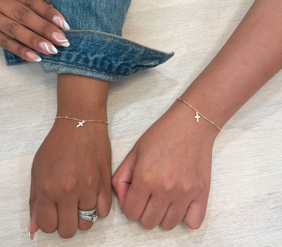 Two women's wrists on a light wood table wearing matching delicate gold chain bracelets with small cross charms; one hand has white French-tip nails, silver rings and a rolled denim cuff.