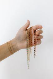 Close-up of a hand holding delicate gold chain necklaces with tiny colorful beads, layered gold bracelets on wrist against a minimalist white background