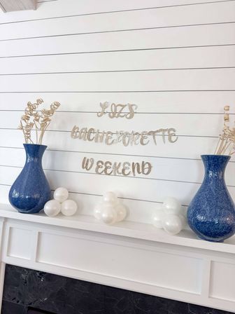 Cheerful bachelorette party mantel with a sparkly "bachelorette weekend" banner on white shiplap, two blue ceramic vases with dried curly stems, and small clusters of white balloons.