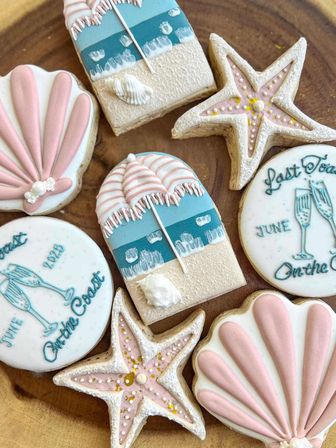Pastel beach-themed decorated sugar cookies on a wooden board: pink scallop shells, starfish with gold sprinkles, teal-and-sand umbrella beach scenes, and round celebration cookies reading ‘June On the Coast’.