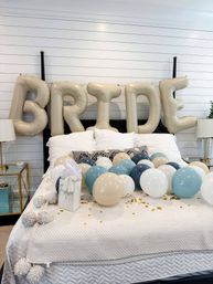 Bedroom styled for a bridal celebration with oversized gold 'BRIDE' letter balloons on a white shiplap wall, beige, blue and white balloons and gold confetti scattered across a white quilted bed.