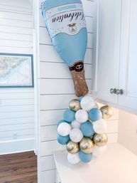 Blue foil champagne bottle balloon mounted on white shiplap wall pouring a cluster of blue, white and metallic gold celebration balloons onto a bright modern farmhouse kitchen countertop