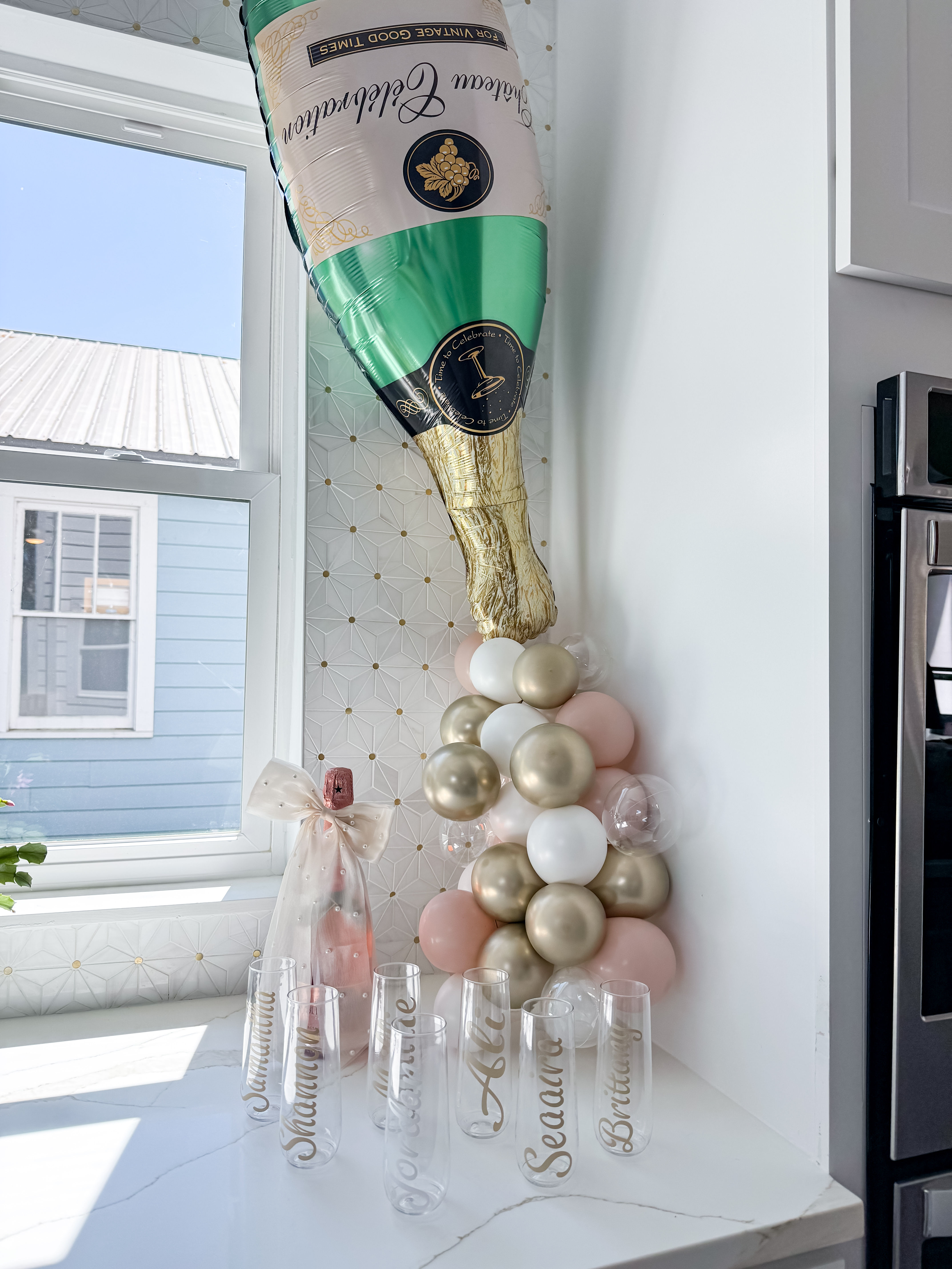 Festive kitchen corner with oversized foil champagne bottle balloon and blush, gold, and white balloon cluster over a marble countertop, wrapped sparkling wine bottle and clear champagne flutes ready for a bubbly celebration