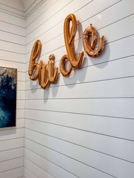 Gold foil balloons spelling "bride" mounted on a white horizontal shiplap wall in an interior corner, with a blue abstract painting partially visible — playful wedding decor.