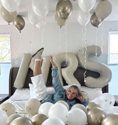 Smiling woman lounging on a bed surrounded by white and gold balloons and oversized beige "MRS" letter balloons in a bright bedroom