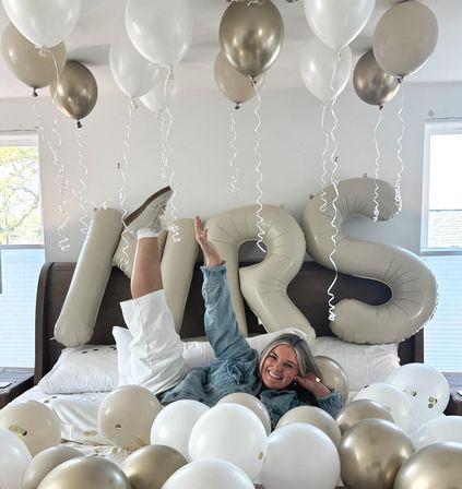 Smiling woman lounging on a bed surrounded by white and gold balloons and oversized beige "MRS" letter balloons in a bright bedroom