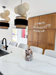 Modern bright kitchen with white marble island, black-and-rattan pendant lights and cream balloons, wooden cabinets with bachelorette weekend signage, and a glass jar of decorative shells on the counter.