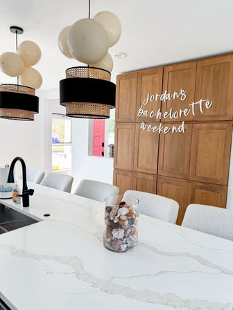 Modern bright kitchen with white marble island, black-and-rattan pendant lights and cream balloons, wooden cabinets with bachelorette weekend signage, and a glass jar of decorative shells on the counter.