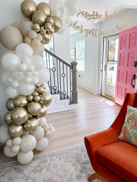 Home entryway decorated with a white and gold balloon garland around a staircase, bright pink front door, glitter birthday banner overhead, and an orange accent chair with patterned pillow.